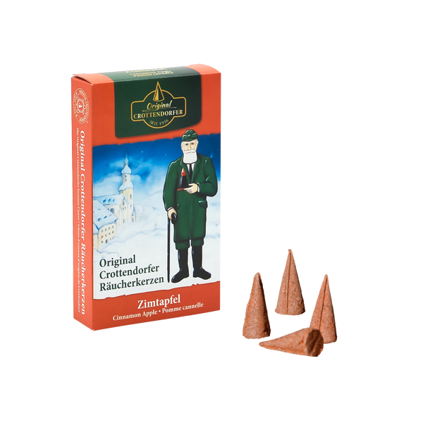 Eine Schachtel Original Crottendorfer Räucherkerzen Zimtapfel steht aufrecht und zeigt eine historische Figur und eine verschneite Dorfszene. Davor sind drei braune Räucherkerzen platziert, die an Bratäpfel erinnern.