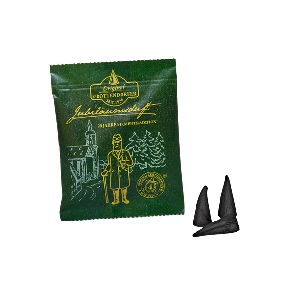 Eine grüne Verpackung mit der Aufschrift „Original Crottendorfer Räucherkerzen“, die mit einer goldenen Illustration verziert und mit der Aufschrift „Räucherkerzen Probierpackung Jubiläumsduft 90 Jahre“ steht auf weißem Hintergrund und spiegelt die erzgebirgische Räuchertradition wider; daneben liegen zwei kegelförmige schwarze Räucherstäbchen.