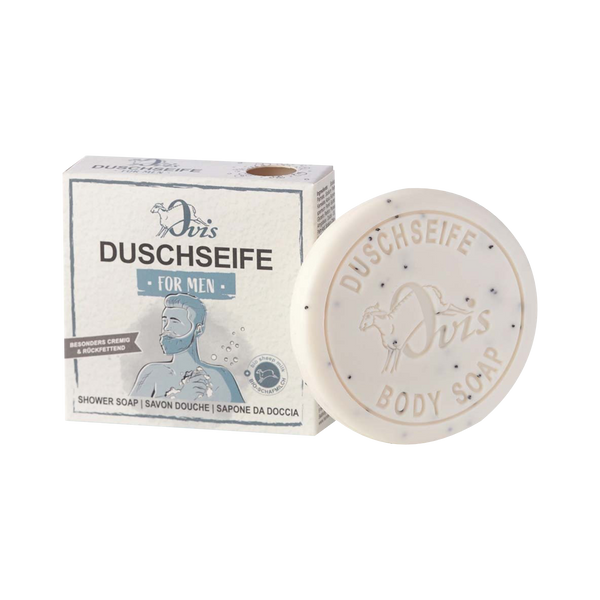 Ein runder Riegel Ovis Duschseife für Männer mit Sheabutter liegt neben der Schachtel, die einen bärtigen Mann beim Duschen zeigt und Text in Deutsch, Französisch, Italienisch und Englisch enthält.