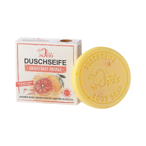 Ein rundes gelbes Stück Ovis Duschseife Grapefruit-Orange liegt neben der Schachtel, die Bilder von Grapefruit und Orange zeigt und den Duft als Grapefruit-Orange kennzeichnet.