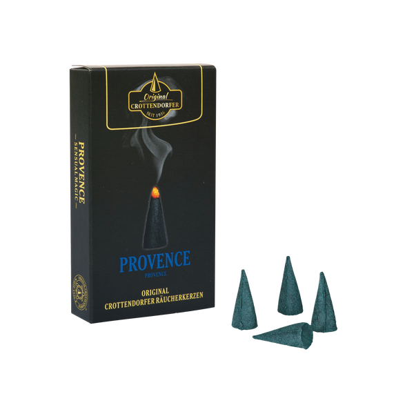 Eine Schachtel mit der Aufschrift „Räucherkerzen Provence“ von Original Crottendorfer Räucherkerzen steht aufrecht neben vier dunkelblauen Räucherkegeln – einer liegt flach, drei stehen aufrecht – auf weißem Hintergrund; jeder verströmt einen romantischen Duft, der von Gartennelke und französischem Honig inspiriert ist.