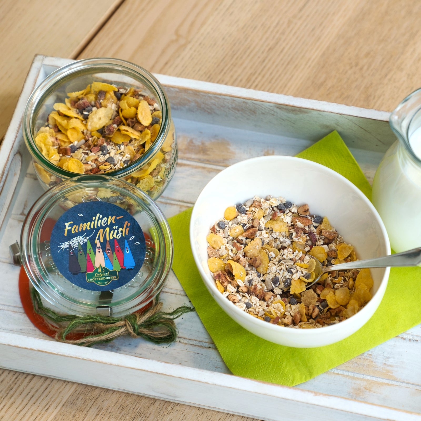Ein Schälchen veganes Hausgemachtes Müsli 200 g von Original Crottendorfer mit Löffel steht auf einer grünen Serviette, daneben ein Glasgefäß und eine Milchflasche auf einem Holztablett, alles auf einem hellen Holztisch.