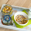 Ein Schälchen veganes Hausgemachtes Müsli 200 g von Original Crottendorfer mit Löffel steht auf einer grünen Serviette, daneben ein Glasgefäß und eine Milchflasche auf einem Holztablett, alles auf einem hellen Holztisch.