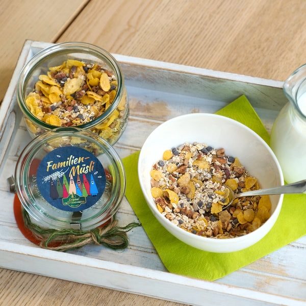 Ein Schälchen veganes Hausgemachtes Müsli 200 g von Original Crottendorfer mit Löffel steht auf einer grünen Serviette, daneben ein Glasgefäß und eine Milchflasche auf einem Holztablett, alles auf einem hellen Holztisch.