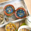 Zwei Gläser mit der Aufschrift Hausgemachtes Müsli 200 g von Original Crottendorfer stehen auf einem Holztablett neben Schüsseln mit Müsli und einem Glaskrug mit Milch auf dem Tisch.