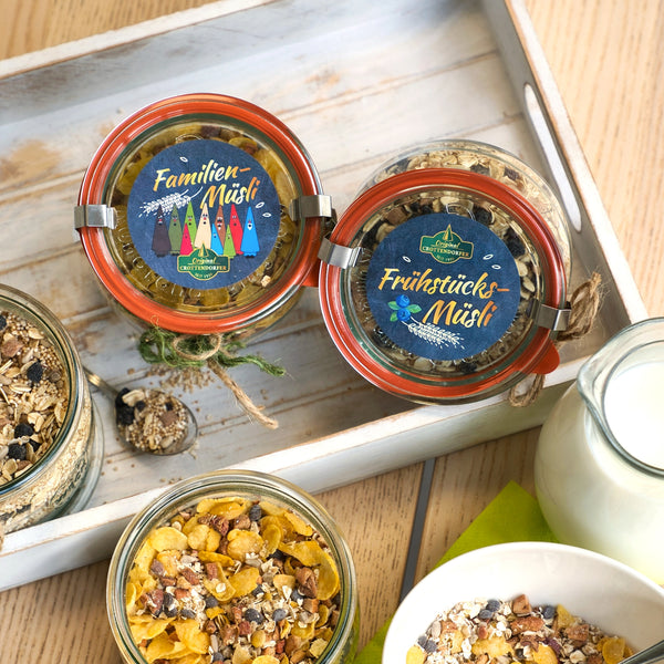 Zwei Gläser mit der Aufschrift Hausgemachtes Müsli 200 g von Original Crottendorfer stehen auf einem Holztablett neben Schüsseln mit Müsli und einem Glaskrug mit Milch auf dem Tisch.