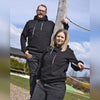 Zwei Personen in passenden Original Crottendorfer Kapuzenpullovern stehen draußen an einem Holzpfosten mit Seil. Der leicht bewölkte Himmel, Grünflächen und ein Gebäude bilden die Kulisse für ihren entspannten Streetwear-Look.