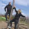 Zwei Personen im Kapuzenpullover „Hoodie Crottendorfer Räucherkerzen“ von Original Crottendorfer präsentieren sich auf einem Outdoor-Spielplatz im Streetwear-Look. Einer steht auf Holzbalken, während der andere sich an ihn lehnt und die Arme ausstreckt. Im Hintergrund eine üppige Grasfläche, Bäume und ein wolkenloser blauer Himmel.