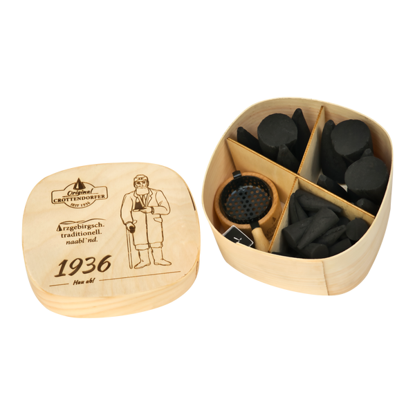 Die „Spanschachtel 1936“ von Original Crottendorfer Räucherkerzen ist eine Holzschachtel mit einem Deckel, auf dem eine Figur und ein Text eingraviert sind. Sie verfügt über Fächer, die mit schwarzen, zylindrischen und unregelmäßig geformten Holzkohlestücken gefüllt sind, sowie über ein kleines rundes Metallgitter – was sie zu einem einzigartigen Geschenkset mit Räucherzubehör macht.
