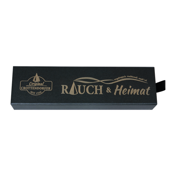 Eine rechteckige schwarze Schubbox „RAUCH & Heimat“ von Original Crottendorfer Räucherkerzen mit goldener Beschriftung: „Original Crottendorfer“, „RAUCH & Heimat“ und „erzgebirgisch. traditionell. rauch’ mal.“ Die Schachtel eignet sich ideal als Geschenk oder zur Aufbewahrung Ihrer Lieblings-Räucheraccessoires und verfügt auf der rechten Seite über eine kleine schwarze Zuglasche.