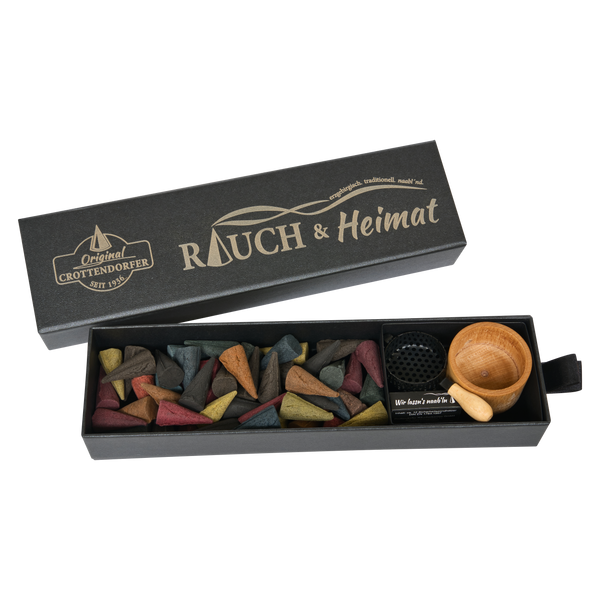 Eine Schubbox „RAUCH & Heimat“ von Original Crottendorfer Räucherkerzen enthält bunte Räucherkegel, eine kleine Holzschale und einen Metallhalter – stilvolle Räucheraccessoires –, die ordentlich in der offenen schwarzen Schachtel angeordnet sind und sie so zum perfekten Geschenk machen.