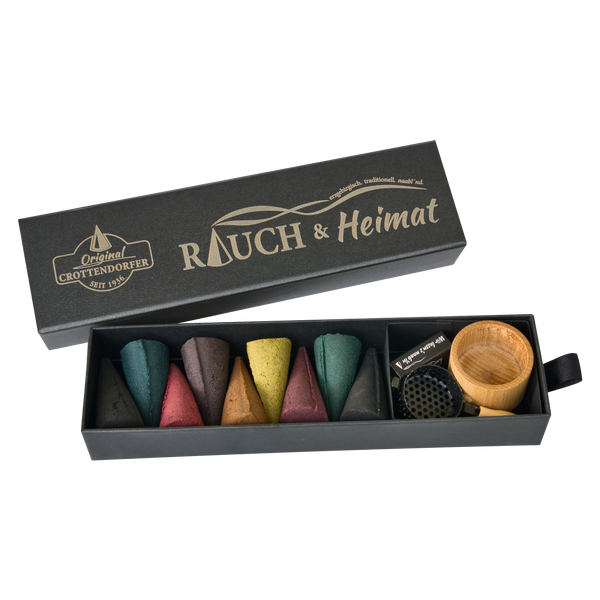 Die Schubbox „RAUCH & Heimat“ von Original Crottendorfer Räucherkerzen enthält farbenfrohe Räucherkegel, einen kleinen Holzhalter und ein Metallgitter – alles unverzichtbare Räucheraccessoires –, die geschmackvoll in der stilvollen Schubbox angeordnet sind, was sie zu einem perfekten Geschenk für jeden Anlass macht.