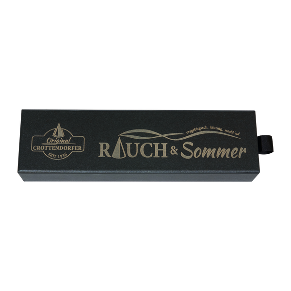 Die Schubbox „RAUCH & Sommer“ von Original Crottendorfer Räucherkerzen ist eine rechteckige schwarze Schachtel mit goldfarbener Beschriftung, auf der die Aufschriften „Rauch & Sommer“ und „Original Crottendorfer Seit 1936“ sowie die Abbildung eines kleinen Segelboots zu sehen sind. Sie ist für Räucherkerzen konzipiert, verfügt auf der rechten Seite über eine schwarze Aufreißlasche und bietet eine klassische Lösung zum Mückenschutz.