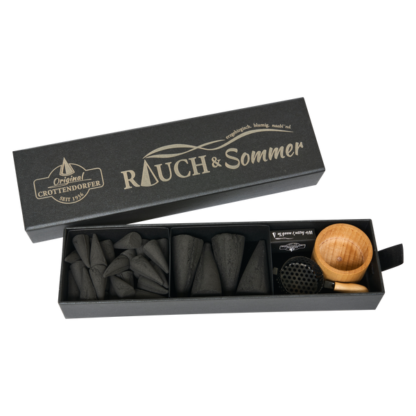 Die Schubbox „RAUCH & Sommer“ von Original Crottendorfer Räucherkerzen ist eine elegante, rechteckige, schwarze Geschenkbox mit dezentem „Rauch & Sommer“-Logo. Im Inneren, unter dem teilweise geöffneten Deckel, befinden sich kegelförmige Räucherkegel zum Mückenschutz, eine kleine Holzschale und ein Halter aus Metallgitter.