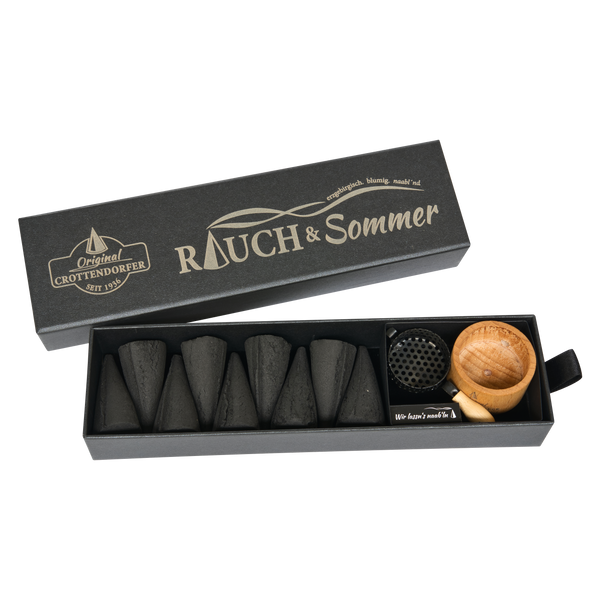 Eine Schubbox „RAUCH & Sommer“ von Original Crottendorfer Räucherkerzen mit sechs schwarzen Räucherkegeln, einem Metallhalter und einer kleinen Holzschale ist vor einem weißen Hintergrund ansprechend in Szene gesetzt.