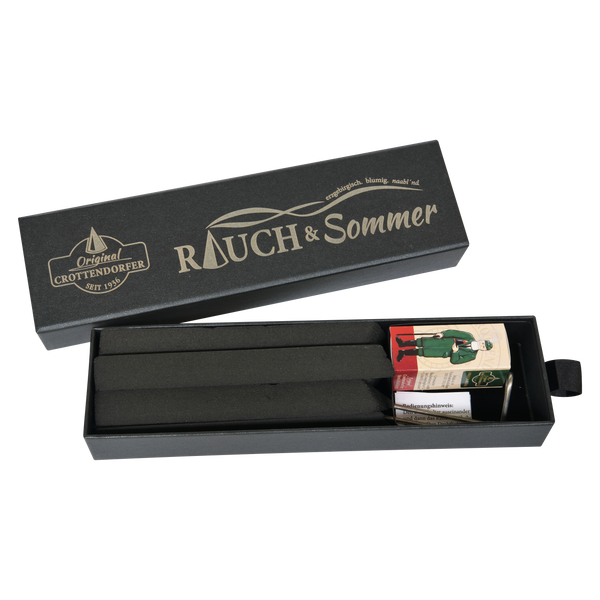 Eine offene, schwarze, rechteckige Schubbox von RAUCH & Sommer aus dem Sortiment von Original Crottendorfer Räucherkerzen enthält zwei lange schwarze Stäbchen und eine kleine Streichholzschachtel, auf der eine Person in grüner Kleidung abgebildet ist.