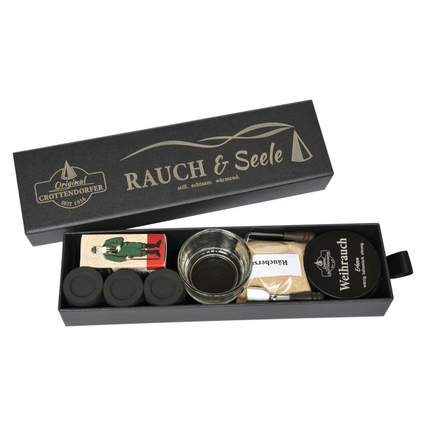 Eine rechteckige schwarze Schubbox „RAUCH & Seele“ von Original Crottendorfer Räucherkerzen enthält ein Geschenkset mit Räucherutensilien, bestehend aus Räucherkegeln, einem Metallhalter, einer Holzpinzette, Streichhölzern und einer runden Dose mit der Aufschrift „Weihrauch Extra“. Die Artikel sind ordentlich in der offenen Schachtel angeordnet.