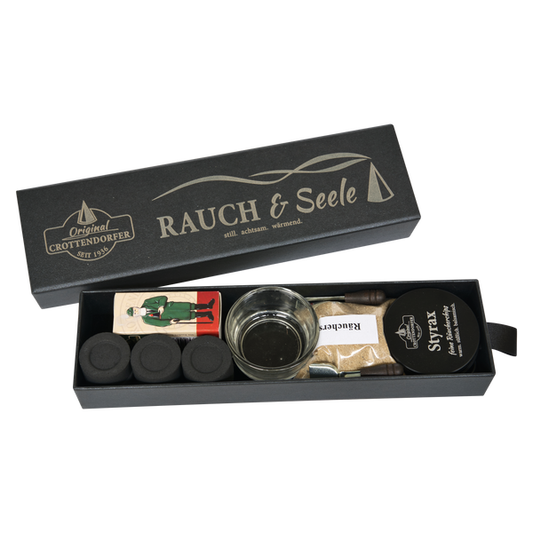 Eine schicke schwarze „Schubbox RAUCH & Seele“ von Original Crottendorfer Räucherkerzen enthält eine sorgfältig zusammengestellte Auswahl an Räucherzubehör, darunter Räucherkegel, einen Glashalter, eine Holzfigur, Streichhölzer, einen Metallhalter und eine Packung mit der Aufschrift „St. Felix“ – alles wunderschön arrangiert in einer eleganten Geschenkbox vor weißem Hintergrund.