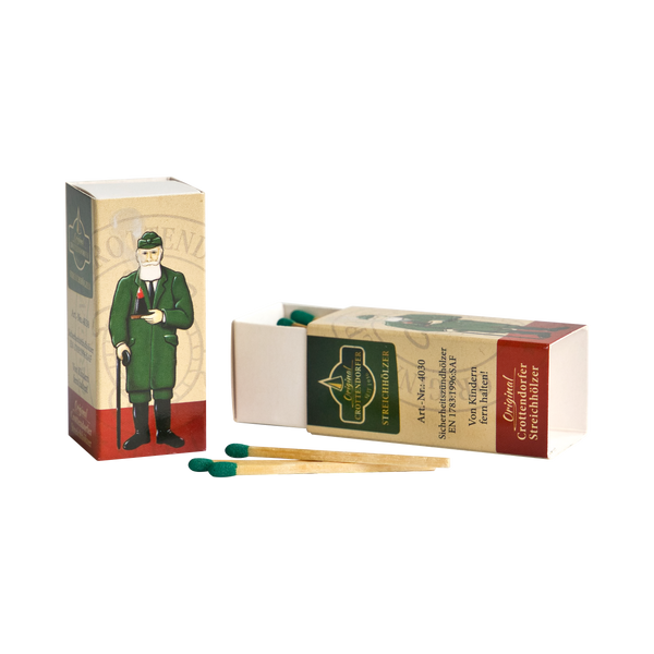 Zu sehen sind zwei Streichholzschachteln der Marke „Original Crottendorfer“ – im klassischen Design mit der Abbildung eines Mannes in grüner Uniform; eine Schachtel steht aufrecht, während die andere offen liegt und den Blick auf die darin teilweise sichtbaren Sicherheitsstreichhölzer mit grünen Spitzen freigibt, wobei zwei Streichhölzer vorne liegen.