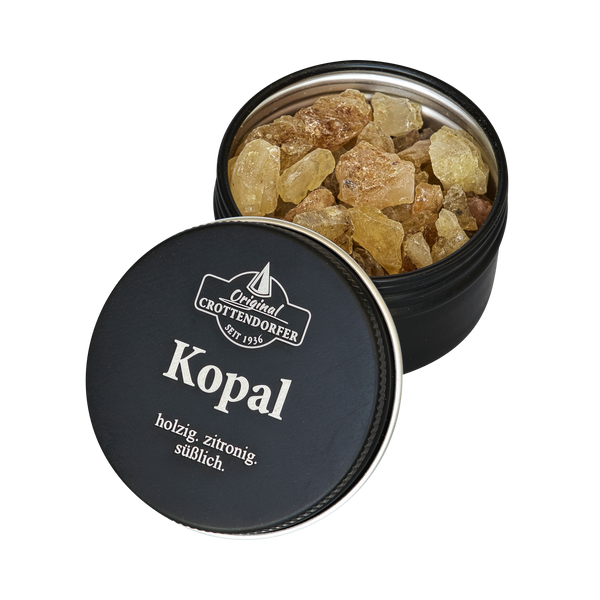 Eine schwarze Blechdose mit der Aufschrift „Kopal in der Schraubdeckeldose“ von Original Crottendorfer Räucherkerzen, deren Deckel einen Spalt offen steht, gibt den Blick auf gelblich-braunes Kopalharz frei, das als holzig, zitronig und süßlich beschrieben wird – ideal für jeden Weihrauchbrenner, der nach reinigenden Eigenschaften sucht.