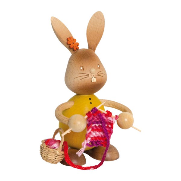 Der Stupsi Hase mit Strickzeug von Drechslerei Kuhnert ist eine handgefertigte Hasenfigur aus Holz, die mit rotem und rosa Garn strickt. Er trägt ein gelbes Hemd, eine orangefarbene Blume am Ohr und hält Stricknadeln, während daneben ein kleiner Korb mit Garnknäueln steht.