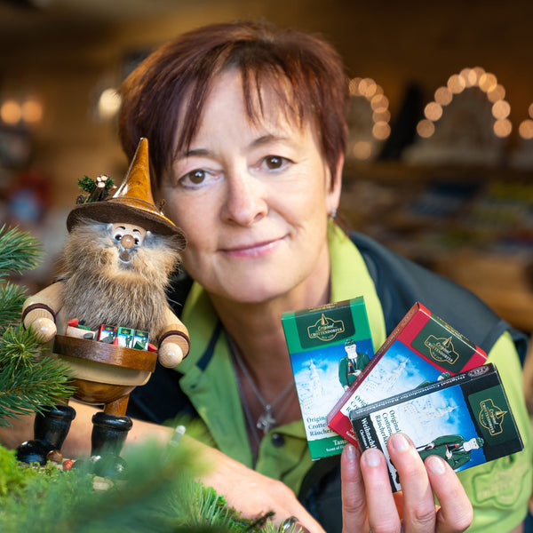 Jemand hält mehrere Schokoladenpackungen neben einer kleinen Figur, dem Original Crottendorfer Räucherkerzenverkäufer der DWU Drechselwerkstatt Uhlig. Der warm beleuchtete Hintergrund ist mit verschwommenen Lichtern dekoriert und fängt den gemütlichen Charme original erzgebirgischer Handwerkskunst ein.