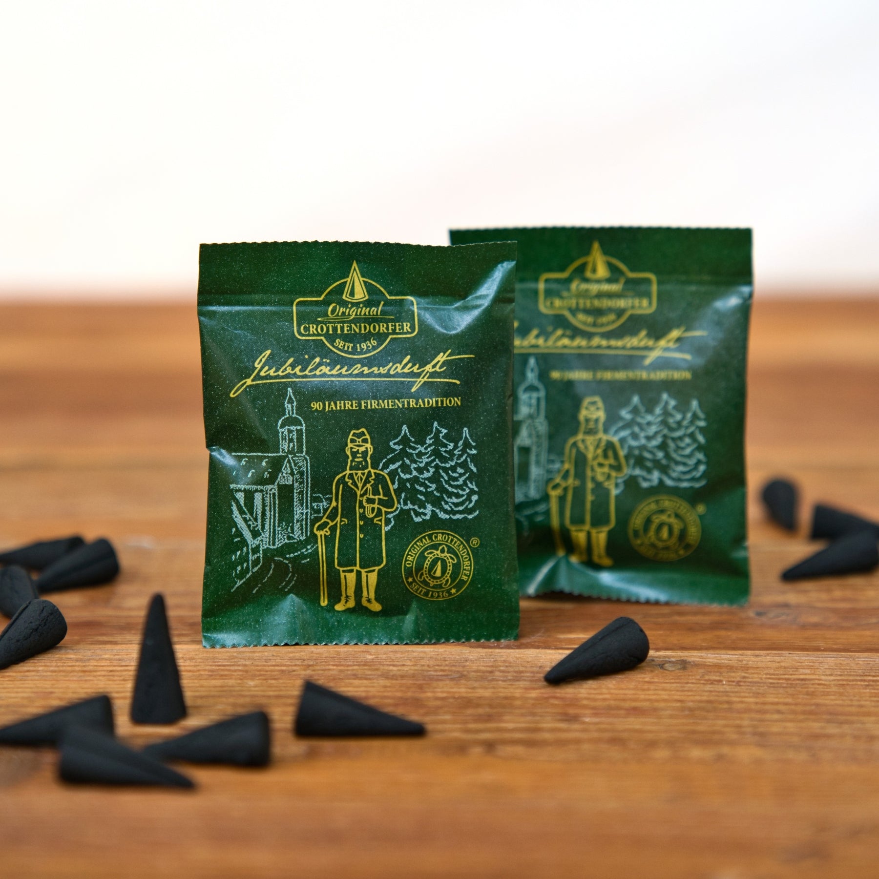 Zwischen mehreren kleinen, schwarzen, kegelförmigen Gegenständen auf einer Holzunterlage stehen zwei grüne Päckchen mit goldenen Illustrationen und der Aufschrift „Original Crottendorfer Räucherkerzen“. Die Päckchen tragen die Aufschrift „Räucherkerzen Probierpackung Jubiläumsduft 90 Jahre“ und zeigen eine Zeichnung eines Mannes und von Bäumen – eine Hommage an die reiche erzgebirgische Räuchertradition.