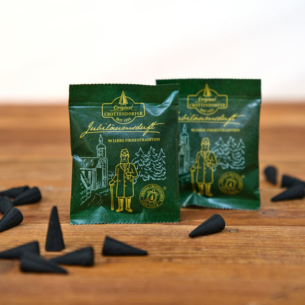 Zwischen mehreren kleinen, schwarzen, kegelförmigen Gegenständen auf einer Holzunterlage stehen zwei grüne Päckchen mit goldenen Illustrationen und der Aufschrift „Original Crottendorfer Räucherkerzen“. Die Päckchen tragen die Aufschrift „Räucherkerzen Probierpackung Jubiläumsduft 90 Jahre“ und zeigen eine Zeichnung eines Mannes und von Bäumen – eine Hommage an die reiche erzgebirgische Räuchertradition.