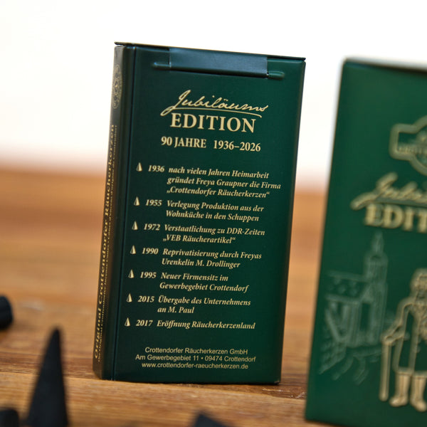 Eine grüne Blechdose – Jubiläumsausgabe „90 Jahre“ von Original Crottendorfer, mit goldenem deutschen Text, der wichtige Meilensteine zur Feier des 90-jährigen Bestehens der Marke (1936–2026) hervorhebt und die Erzgebirgische Tradition würdigt, mit einer ähnlichen Gedenkdose, die im Vordergrund leicht unscharf dargestellt ist.