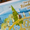 Eine fröhliche grüne Weihnachtsbaum-Zeichentrickfigur zündet vor einer verschneiten Kulisse eine Kerze am „Räucherkerzen-Adventskalender 2026 – Querformat“ von Original Crottendorfer Räucherkerzen an. Die nummerierten Türchen – darunter die Nummern 21 und 24 – fangen die festliche Stimmung der erzgebirgischen Traditionen ein, während die dezente Präsenz der Räucherkerzen zur warmen und gemütlichen Weihnachtsstimmung beiträgt.