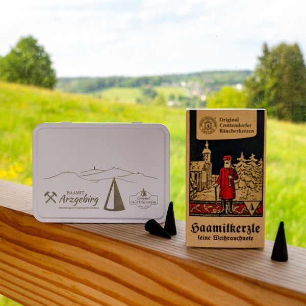 Die Original Crottendorfer Aufbewahrungsbox mit Scharnierdeckel und Lasergravur steht zusammen mit der Räucherkegel-Box und zwei schwarzen Kegeln auf einem Holzsockel. Durch das Fenster im Hintergrund sieht man eine grüne Landschaft.