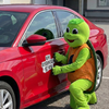 Eine Person in einem grünen Schildkrötenkostüm kniet neben einem roten Auto und klebt einen Original Crottendorfer-Aufkleber mit der Aufschrift "Macht Raacherkerzln, nicht Krieg" auf die Fahrertür.