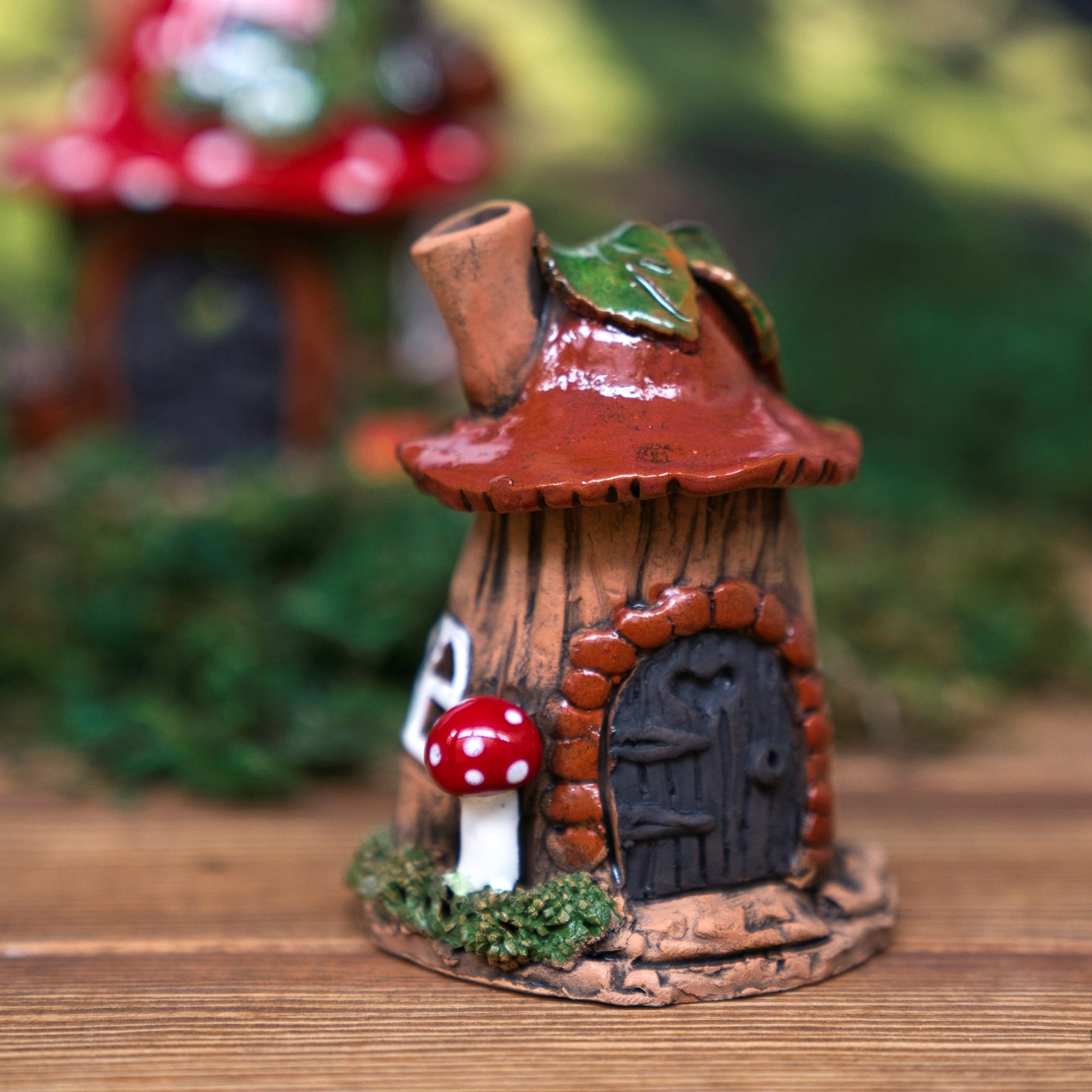 Ein verspieltes „Räucherhaus Keramik Blattpilzhaus“ von Original Crottendorfer in Form eines Baumstamms mit einem roten, pilzartigen Dach, einer gewölbten Tür, einem winzigen Fenster und einer roten Pilzdekoration am Sockel steht auf einer Holzunterlage, umgeben von Grünpflanzen. Dieses bezaubernde Keramikstück ist in drei Farben erhältlich und verleiht jedem Raum Charme und Fantasie.
