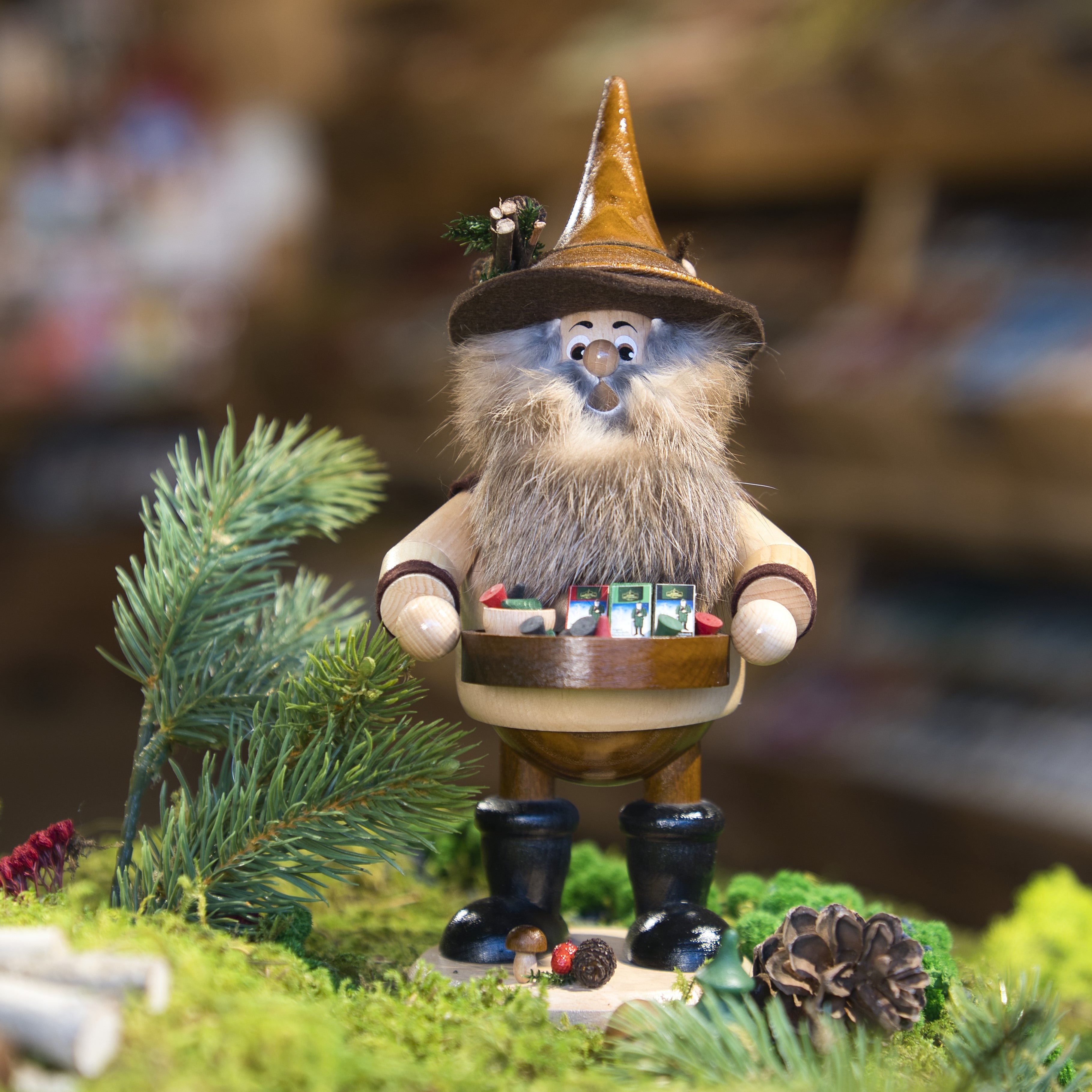 Eine bezaubernde Szene zeigt den Original Crottendorfer Räucherkerzenverkäufer, eine hölzerne Zwergenfigur mit Bart und Zipfelmütze, die auf moosbewachsenem Boden steht. Sie hält ein Tablett mit verschiedenen Gegenständen und ist von Tannenzapfen und immergrünen Zweigen umgeben. Sie ist im klassischen Stil der DWU Drechselwerkstatt Uhlig gefertigt.