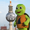 Eine Person in einem großen grünen Schildkrötenkostüm steht vor dem Berliner Fernsehturm, der mit dem Aufkleber "Macht Raacherkerzln, keinen Krieg!" von Original Crottendorfer versehen ist - eine spielerische Anspielung auf erzgebirgische Traditionen unter freiem Himmel.