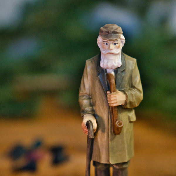 Die Figur "Crottendorfer Räuchermann" von Original Crottendorfer Räucherkerzen zeigt einen älteren Mann mit weißem Bart, Mütze und langem Mantel, der eine Pfeife und einen Spazierstock hält und vor einem unscharfen Hintergrund posiert.