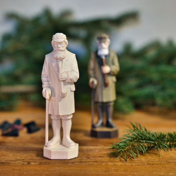 Zwei Figuren "Crottendorfer Räuchermann" von Original Crottendorfer Räucherkerzen, eine helle und eine dunkle, stehen mit Stöcken auf Holz inmitten von Tannenzweigen - eine charmante Anspielung auf traditionelle Handwerkskunst.