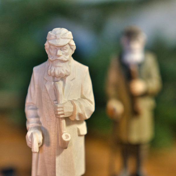 Eine Nahaufnahme der unvollendeten Figur "Crottendorfer Räuchermann" von Original Crottendorfer Räucherkerzen zeigt einen bärtigen Mann mit einer Pfeife. Im Hintergrund ist eine gemalte Version der gleichen Figur zu sehen, die jedoch unscharf ist.