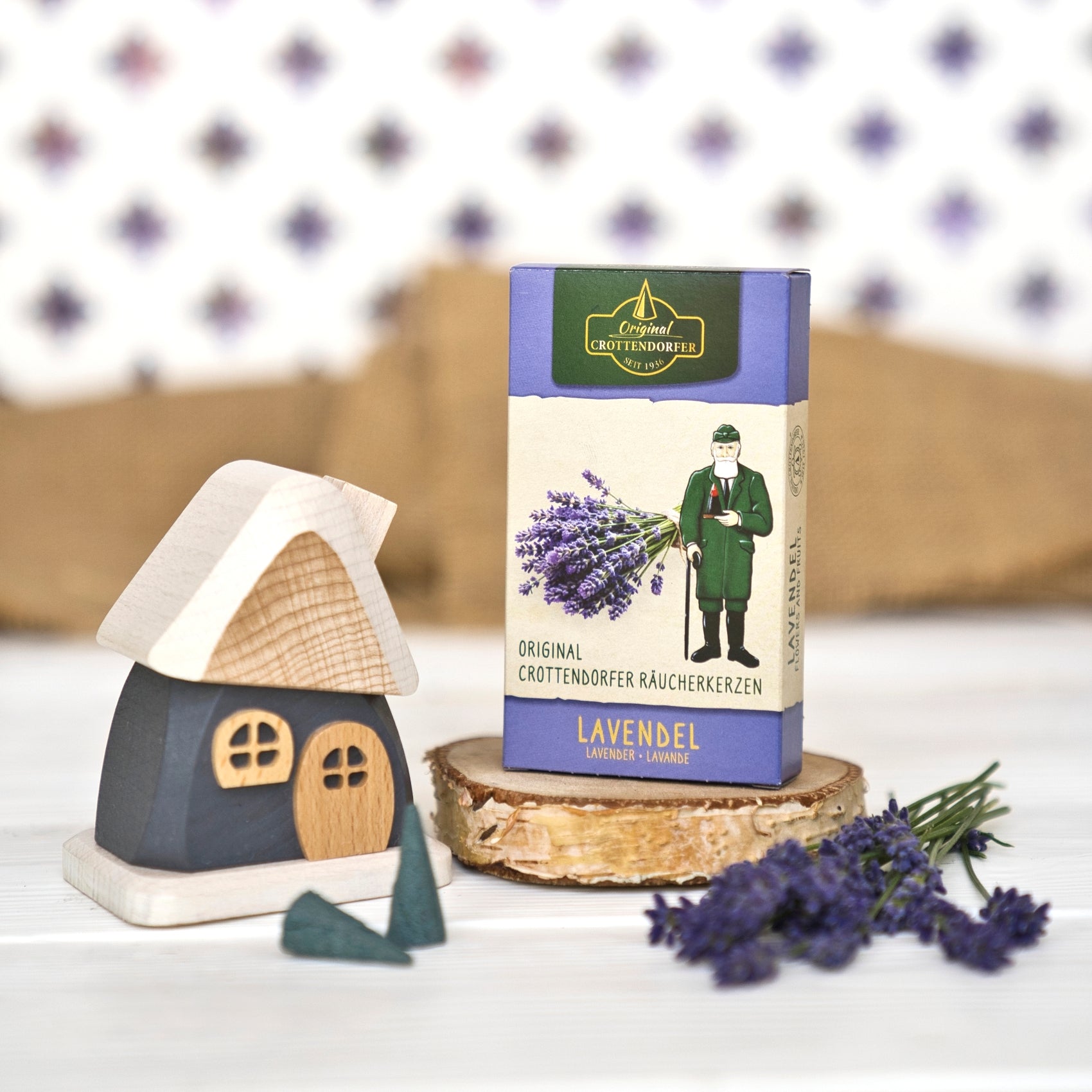Eine Schachtel mit Original Crottendorfer Räucherkerzen „Lavendel“ steht auf einer Holzscheibe neben einem kleinen Holzhäuschen und einem Lavendelzweig und verströmt den blumigen Duft der Provence; im Hintergrund sind eine weiß gemusterte Fläche und ein Stück Sackleinen zu sehen.