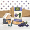 Eine Schachtel mit Original Crottendorfer Räucherkerzen „Lavendel“ steht auf einer Holzscheibe neben einem kleinen Holzhäuschen und einem Lavendelzweig und verströmt den blumigen Duft der Provence; im Hintergrund sind eine weiß gemusterte Fläche und ein Stück Sackleinen zu sehen.