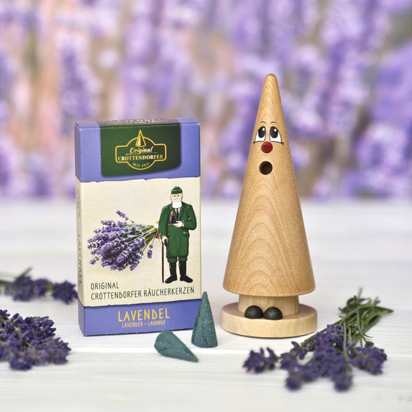 Neben einer Schachtel mit „Original Crottendorfer Räucherkerzen Lavendel“, zwei Räucherkegeln und Lavendelzweigen steht ein Räucherstäbchenhalter in Form eines Holzkegels mit einem überraschten Gesicht. Das Ganze ist vor einem sanft unscharfen Hintergrund angeordnet, der an den blumigen Duft der Provence erinnert.