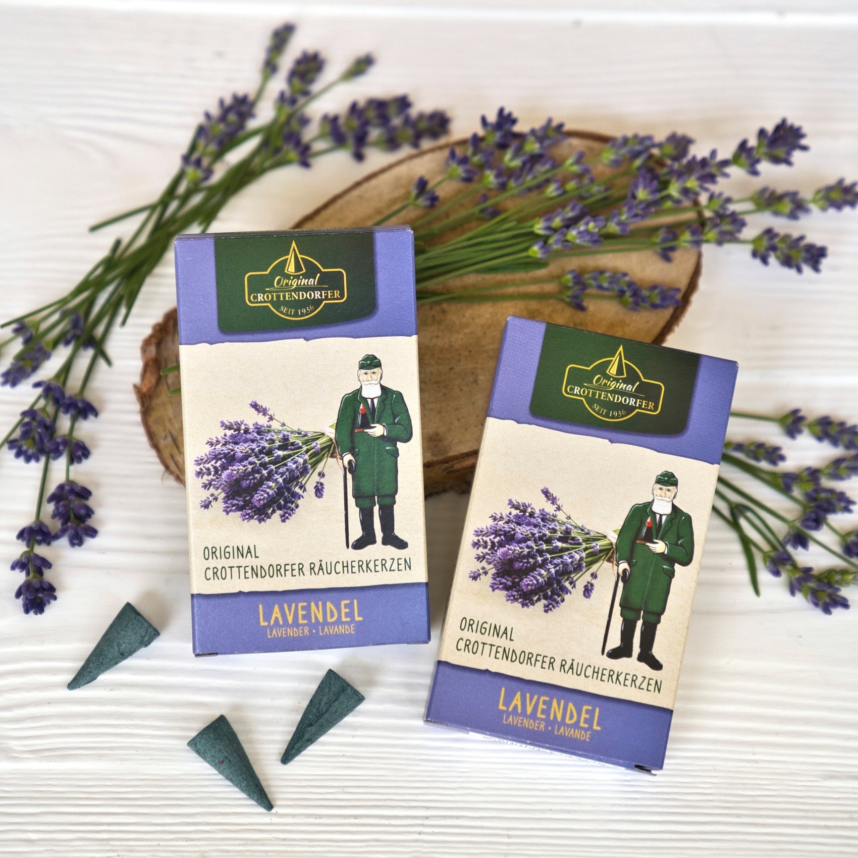 Zwei Schachteln mit Original Crottendorfer Räucherkerzen „Lavendel“, ergänzt durch eine Figur, die Lavendel in der Hand hält, drei Räucherkegel und frische Lavendelzweige, die auf einer weißen Holzunterlage arrangiert sind, versetzen den Betrachter in den blumigen Duft der Provence.