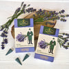 Zwei Schachteln mit Original Crottendorfer Räucherkerzen „Lavendel“, ergänzt durch eine Figur, die Lavendel in der Hand hält, drei Räucherkegel und frische Lavendelzweige, die auf einer weißen Holzunterlage arrangiert sind, versetzen den Betrachter in den blumigen Duft der Provence.