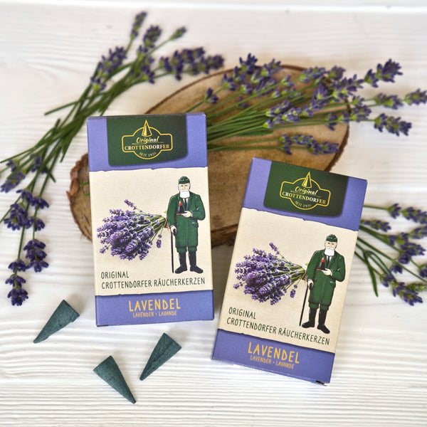 Zwei Schachteln mit Original Crottendorfer Räucherkerzen „Lavendel“, ergänzt durch eine Figur, die Lavendel in der Hand hält, drei Räucherkegel und frische Lavendelzweige, die auf einer weißen Holzunterlage arrangiert sind, versetzen den Betrachter in den blumigen Duft der Provence.