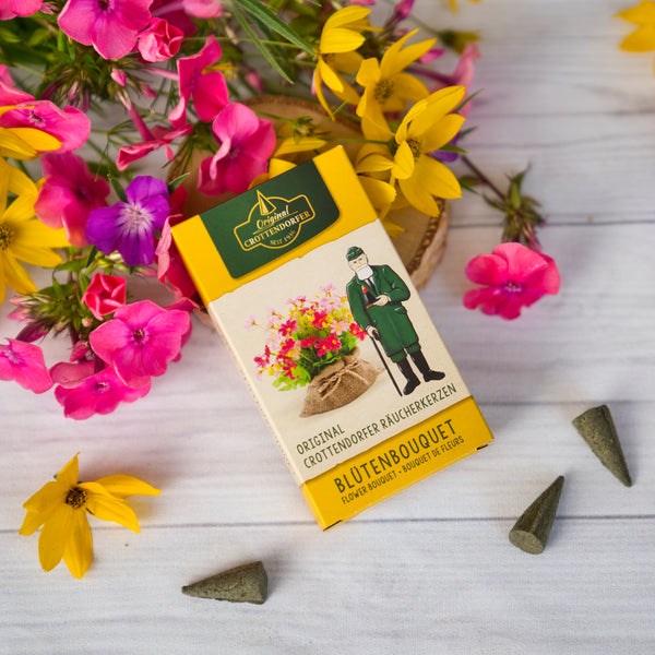 Eine gelbe Schachtel Original Crottendorfer Räucherkerzen Blütenbouquet liegt auf einer weißen Holzfläche zwischen rosa und gelben Blumen und drei Räucherkegeln und verbreitet einen zarten Duft von Jasmin und Rosenblüten.
