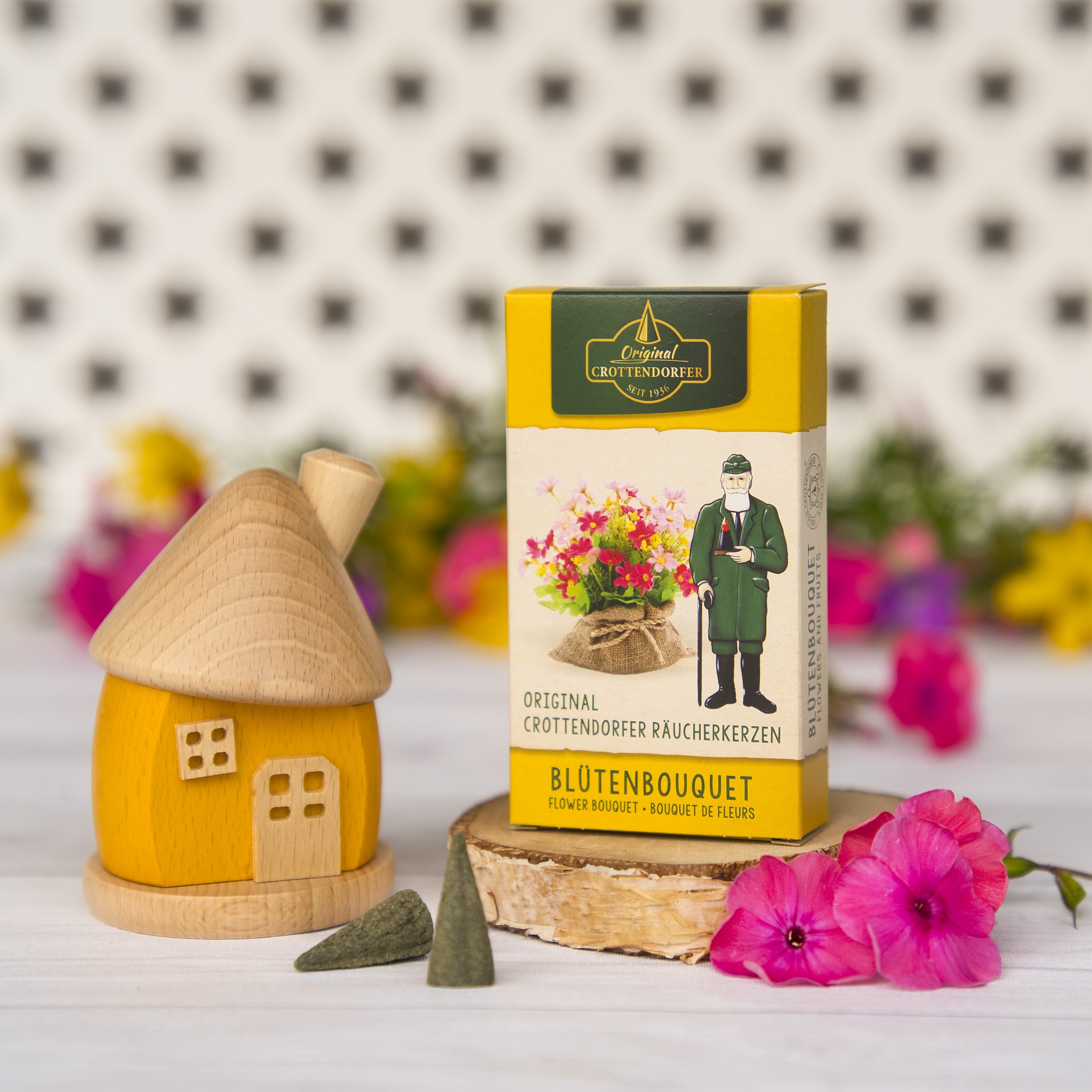 Eine Schachtel mit Original Crottendorfer Räucherkerzen Blütenbouquet steht neben einem kleinen Holzhaus, Blumen und losen Kegeln auf einer weißen Fläche mit floralen Mustern und schafft eine Atmosphäre von sanftem Duft.