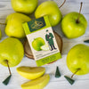 Eine Schachtel Original Crottendorfer Räucherkerzen Grüner Apfel wird mit grünen Äpfeln, Apfelscheiben und grünen Räucherkegeln auf einer weißen Holzfläche präsentiert und verströmt ein frisches Apfelblütenaroma.