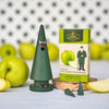 Ein grüner Räucherkegel mit einem überraschten Gesicht steht neben einer Schachtel Original Crottendorfer Räucherkerzen Grüner Apfel, daneben liegen Apfelscheiben auf einer hellen Fläche, die den frischen Duft erahnen lassen.