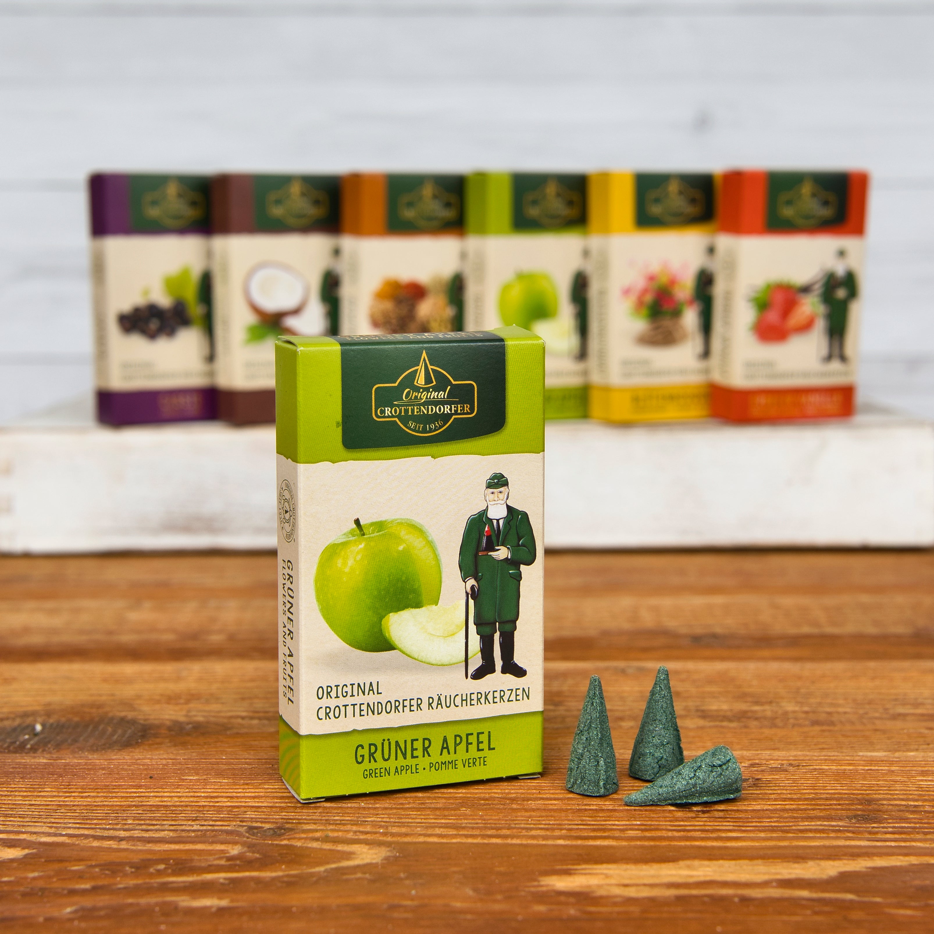 Eine Schachtel Original Crottendorfer Räucherkerzen Grüner Apfel mit grünem Apfelmotiv steht auf Holz. In der Nähe befinden sich drei grüne Räucherkegel, die einen frischen Duft suggerieren. Im Hintergrund erscheinen lebendige, verschwommene Räucherkästchen.