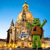 In der Abenddämmerung hält ein Mann im grünen Schildkrötenkostüm ein Schild mit der Aufschrift "Macht Raacherkerzln, nicht Krieg!" - die Botschaft des Original Crottendorfer Aufklebers - und feiert vor einem großen, beleuchteten historischen Gebäude erzgebirgische Tradition.